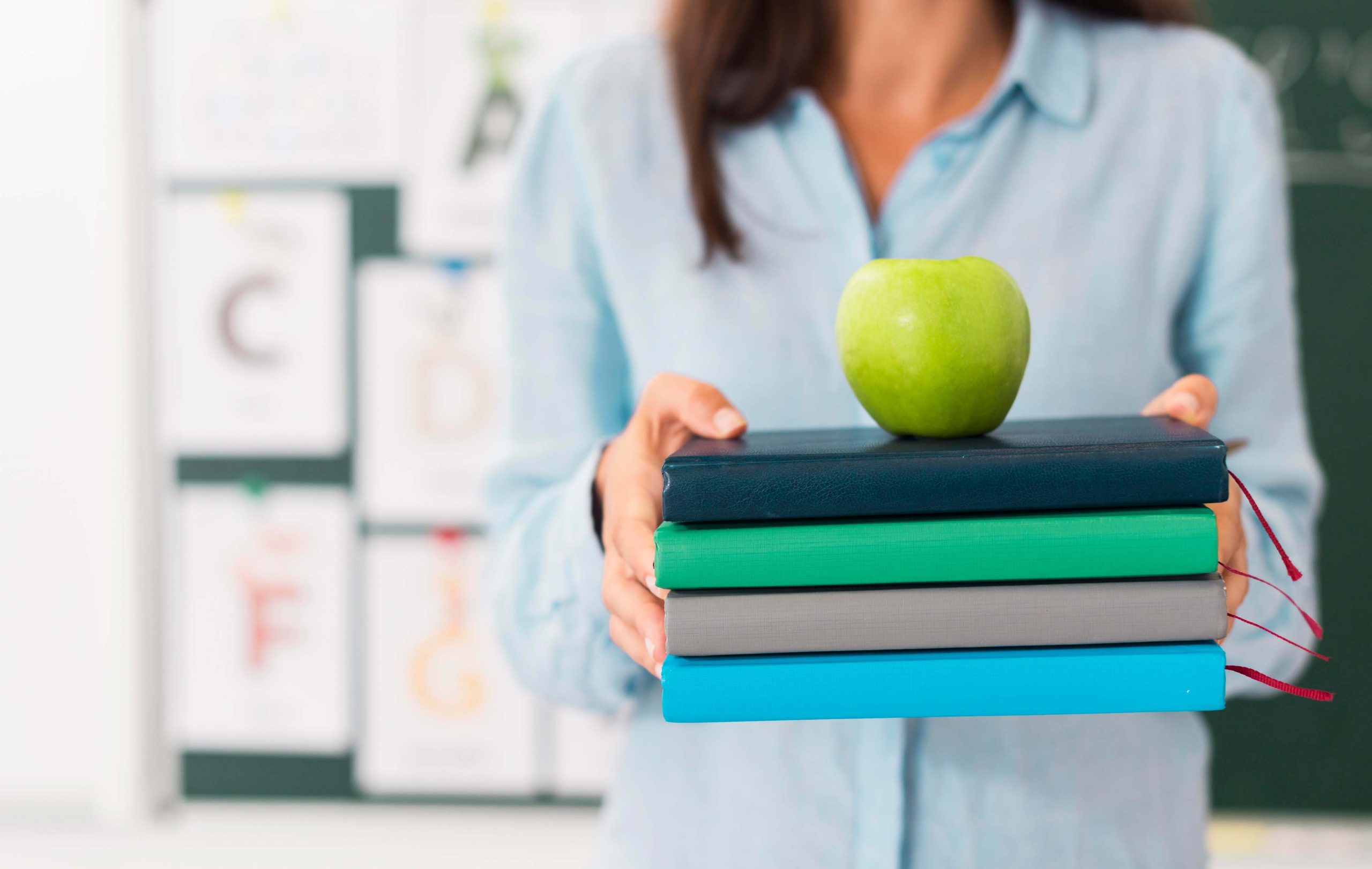 Una persona sosteniendo varios libros con una manzana verde en la parte superior en un aula.
